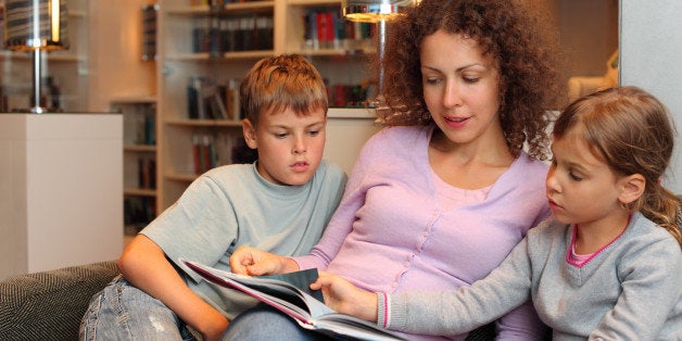 Son and daughter with their mother sit on sofa and read book in room; focus on woman