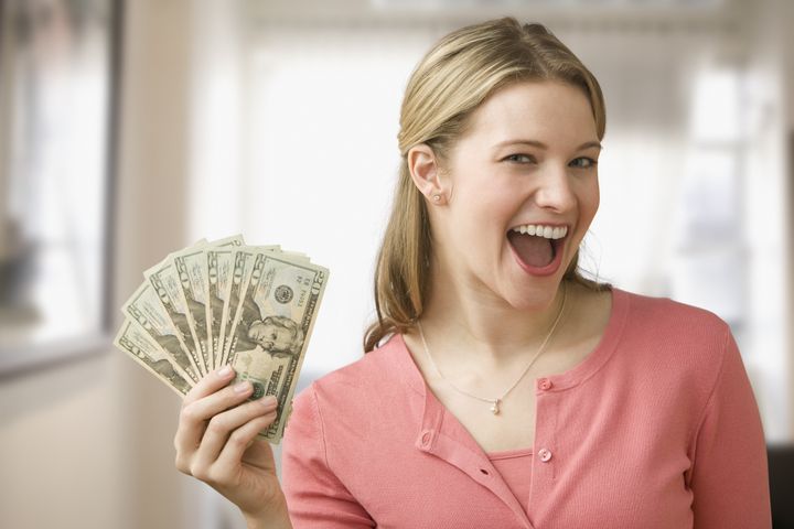 A young woman is holding up cash in a fan and smiling at the camera. Horizontal shot.