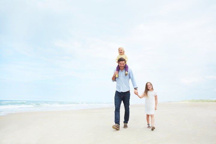 A loving father with a young daughter on his shoulders while holding the other's hand on the beach