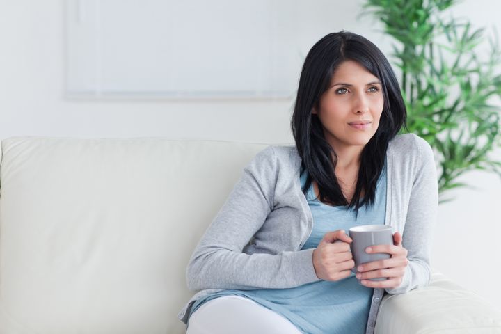 Thinking woman sits on a couch while holding a mug in a living room