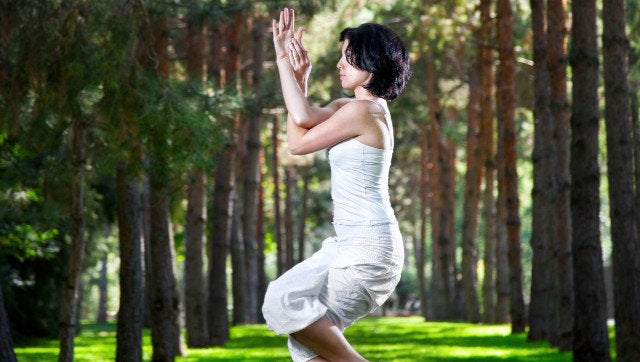 Yoga garudasana eagle pose by woman in white costume on green grass in the park around pine trees