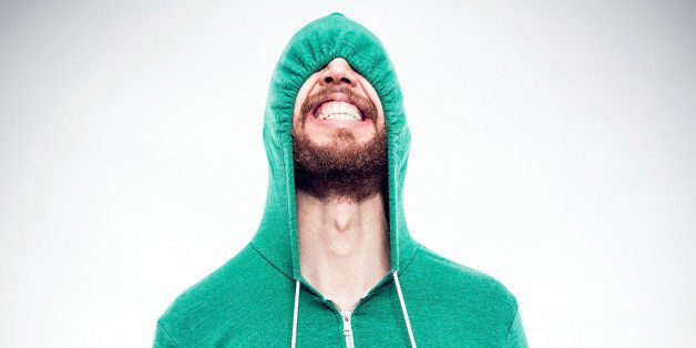 A bearded hipster young man pulls the drawstrings on his green hoodie, partially concealing the funny face he is making, complete with a cheesy grin. Square crop.