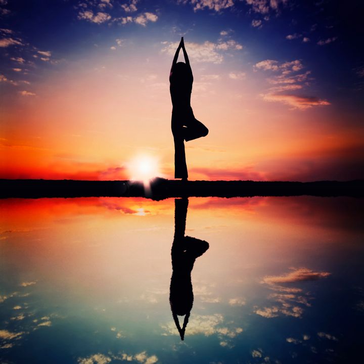 Woman doing tree pose silhouette,silhouette is mirrored below with a sunset sky as backdrop