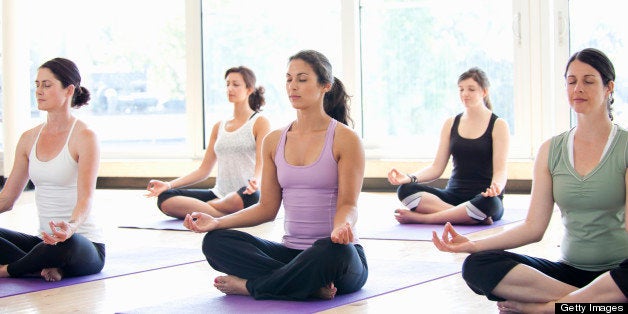 Class of women performing the meditative Sukhasana pose in a bright, modern yoga studio.