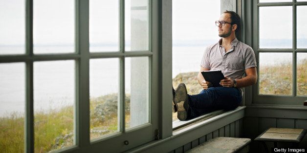Man sitting on window ledge of porch of vacation home looking out holding digital tablet