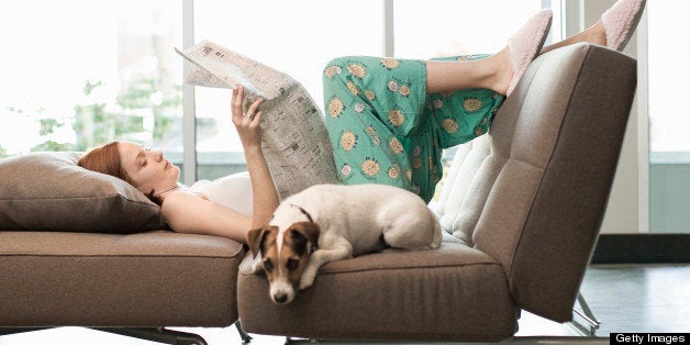 25yr. Cauc woman in pajama bottoms and camisole, laying on couch as she reads newspaper, Jack Russell terrier laying next to her