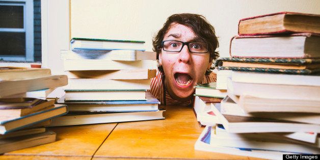 A stressed out college or high school student hiding behind piles of books on top of his desk. He is wearing glasses, and has brown hair.