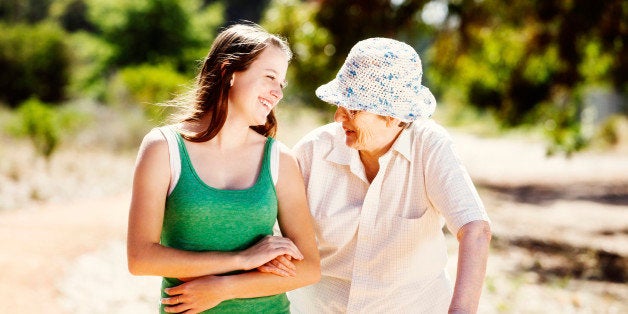 A young woman and an old woman, probably grandmother and granddaughter, with linked arms, chat happily to each other, laughing as they walk through a sunlit forest. 