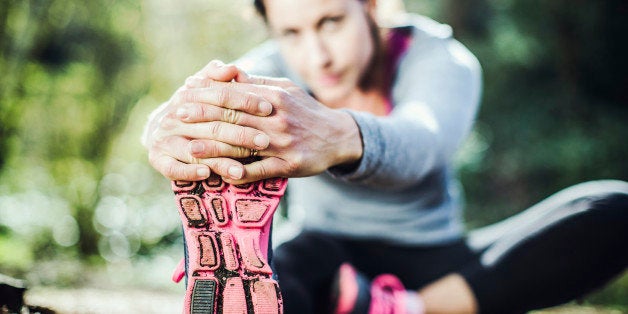 A woman in her early 30's stretches her legs in a bright Pacific Northwest forest after a jog. Selective focus on her shoe and hands in the foreground. Horizontal with copy space.