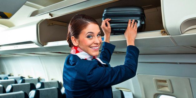 Beautiful flight attendant putting a suitcase in an overhead compartment in an airplane and smiling at the camera.