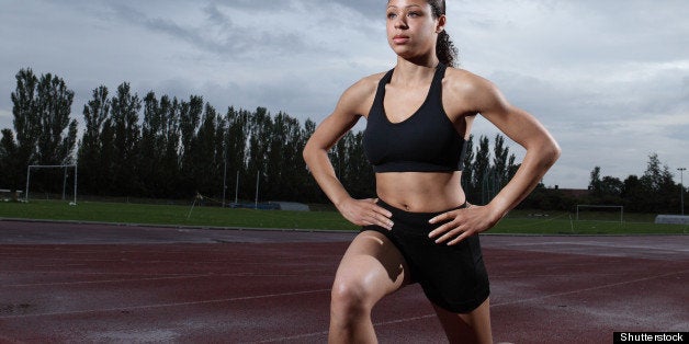 Warm up lunge exercise for quadriceps by fit young female athlete on athletics running track, wearing black lycra sports outfit and running spikes. Grey cloudy sky in background.