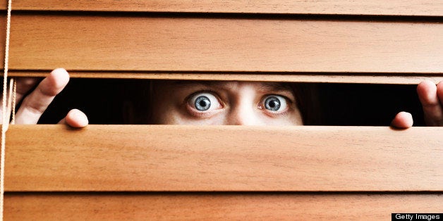 A frightened young woman stares, wide eyed, through the slats of a wooden venetian blind. She could be having home security issues or simply be extremely nervous.