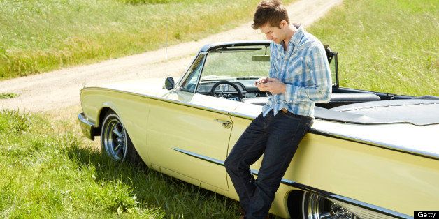 Side view of a young man using a smart phone sending a text message. He is standing by vintage car during summer road trip. Horizontally framed shot. 