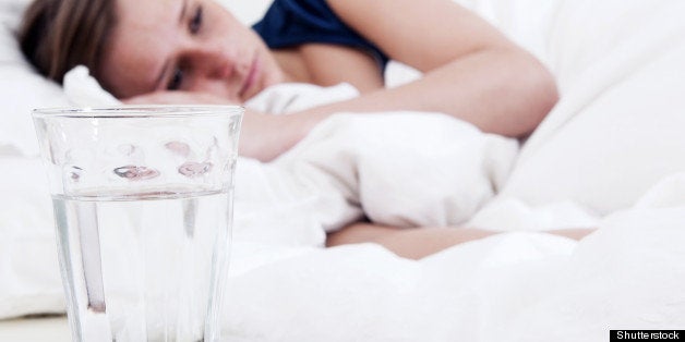 Bedside table with a glass of water, and a blister strip with pills and an awake, sick woman in the background