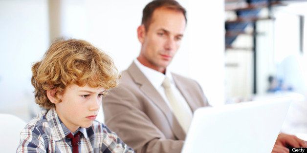 A young boy working on his laptop with his father looking on in the background