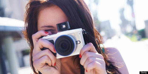 A brunette standing in a urban setting holding a camera in front of her face and taking a picture