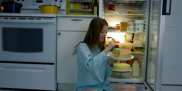 Woman in Pajamas Sits on Kitchen Floor Next to a Fridge Eating a Cake