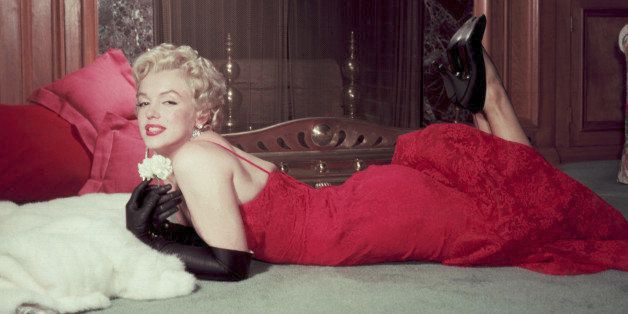1955: EXCLUSIVE Portrait of American actor Marilyn Monroe (1926 - 1962) as she holds a white carnation while lying on her stomach in front of a fireplace. Monroe wears a red evening dress, long black gloves, and black high heels. (Photo by Gene Lester/Getty Images)
