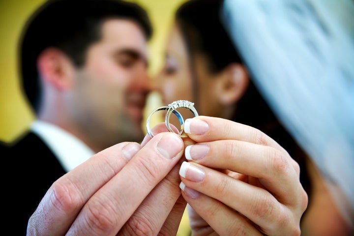 The bride and groom are holding up the wedding rings and kissing in the background. NOTE: this photo has a very shallow depth of field