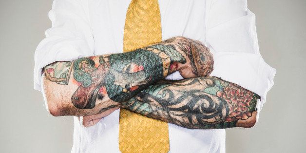 A business man stands with his tattooed arms folded across his white collared shirt and tie. Two forearm sleeve tattoos. Representing a new generation of modern business standards and style.