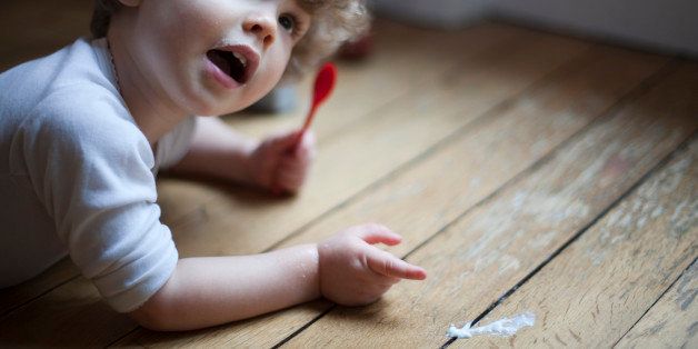 Toddler lying on floor with spoon in hand, pointing to spilled food