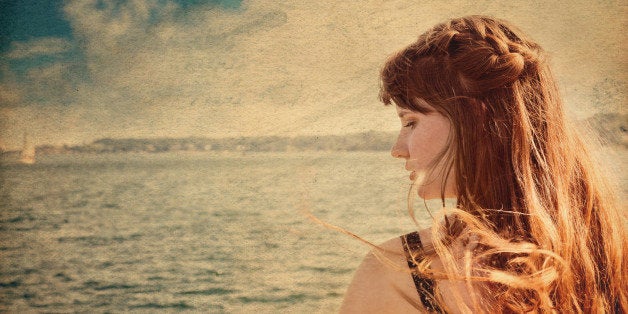 Closeup portrait of young woman in profile with one braid running along top of very windswept hair, looking down with sad expression. Atlantic Ocean and one sailboat are in soft focus in background, overlaid with antique paper texture and wash.