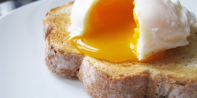 A single poached egg on artisan hand sliced toasted bread. The egg is split to reveal a runny yolk. White dinner plate, non-descript dark wood table and chair just about visible in the background.