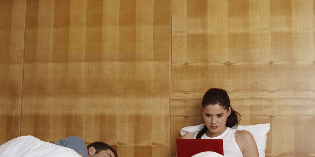 Woman using laptop in bed with sleeping man by her