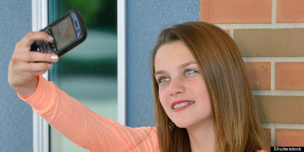 Adorable young girl, with braces, sitting in front of school building with book open on lap but taking photo of self with cell phone camera