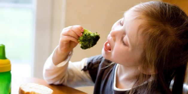 A 4 year old girl sitting at the table enjoying a healthy lunch. She is eating broccoli with her hands, her mouth is wide open. Soft window lighting.