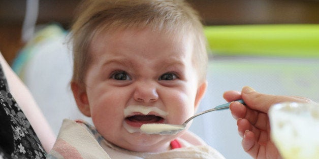 Side view of mother hand feeding frustrated and crying baby girl with spoon.