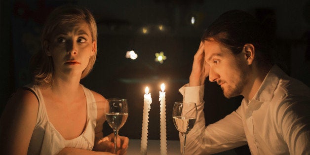 A couple appear to be in a state of tension as if grappling with a serious problem. They are drinking white wine and sit at a table lit by candlelight.