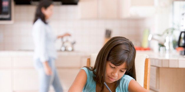 Young girl in kitchen doing homework with mother in background