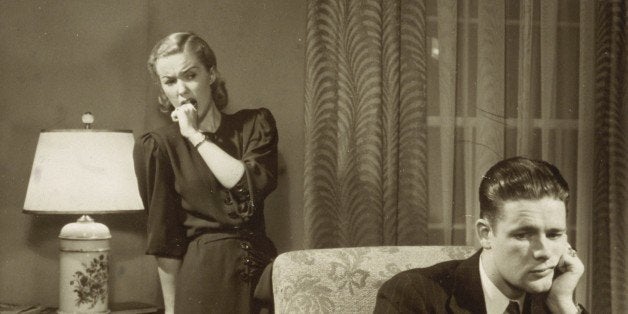 A man sits in an armchair, leaning his chin in one hand and looking depressed, as a woman looks at him with horror, holding her knuckle to her mouth.