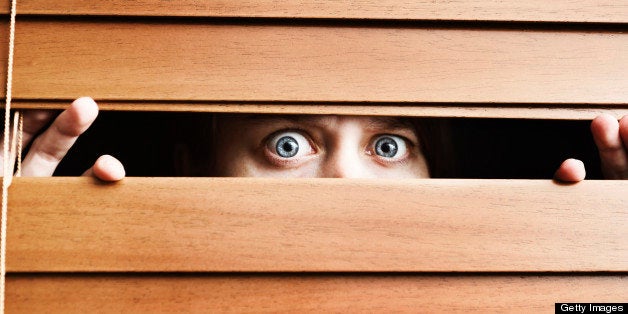 A frightened young woman stares, wide eyed, through the slats of a wooden venetian blind. She could be having home security issues or simply be extremely nervous. 