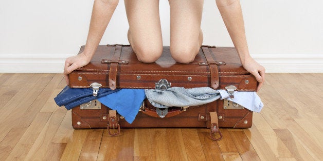 Young woman standing on her knees on overfilled suitcase, trying to close it.