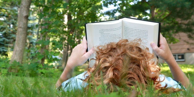 A boy lies on his back in the grass and reads a book.