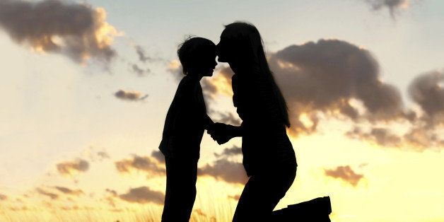 Silhouette of a young mother lovingly kissing her little child on the forehead, outside isolated in front of a sunset in the sky.