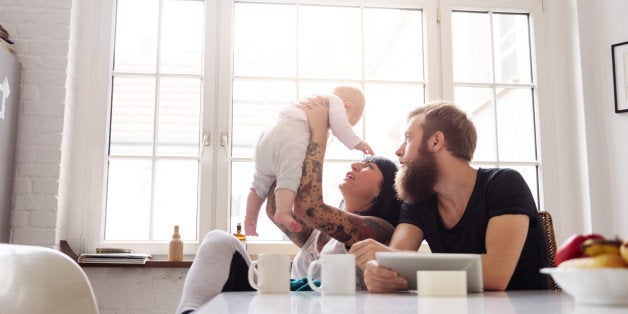 Young tattoed mother and father with newborn baby sitting in their kitchen and having fun together