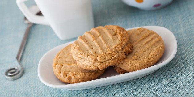 High Key image of 3 peanut butter cookies on a white oblong plate with a blue cloth along with a cup of tea and a polka dot teapot in the background.