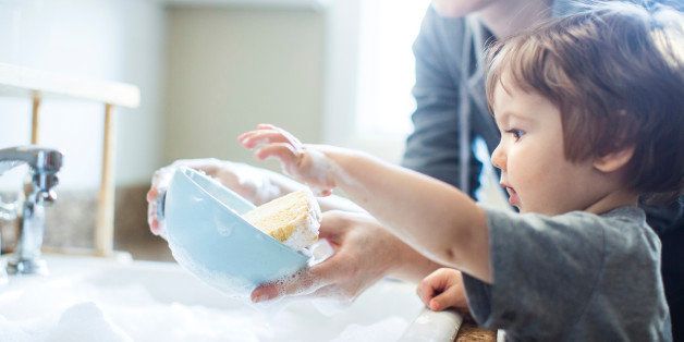 A cute toddler aged boy wearing a blue diaper and tshirt helps out with with washing the dishes, standing on a chair to be able to reach the sink full of soapy bubbles and dishes. His mother smiles behind him as she supervises his sensory perception experience. Bright sunlight comes in through the window behind him, lighting the sparse modern kitchen.
