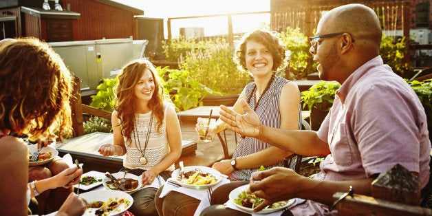 Laughing group of friends sitting together sharing dinner and wine in rooftop garden on summer evening