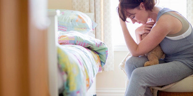 An unhappy woman sits in a child's bedroom with her head in her hands. She is holding a soft toy that belonged to her child. The bed in front of her is empty.