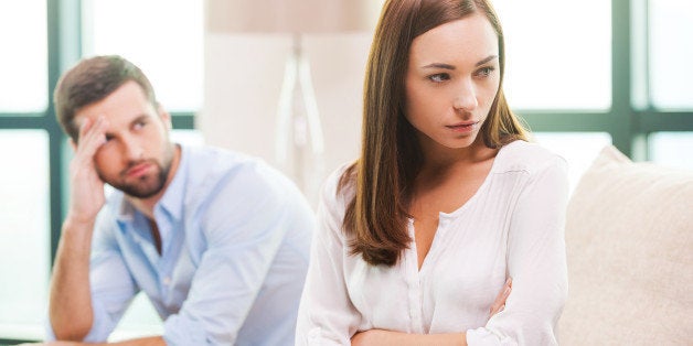 Depressed young woman keeping arms crossed and looking away while man sitting behind her on the couch