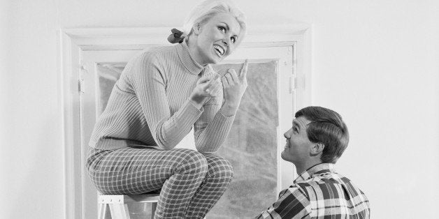 Young woman sitting on step ladder and man standing beside her while painting house