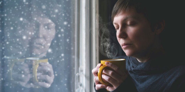 Lonelsome woman drinking cup of coffee by the window of her living room while the snow is falling outside. Selective focus with shallow depth of field.