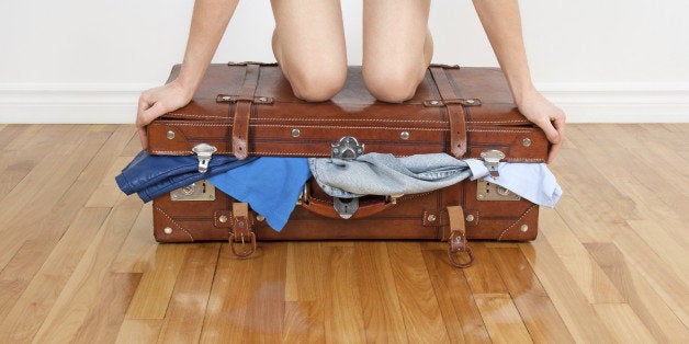 Young woman standing on her knees on overfilled suitcase, trying to close it.