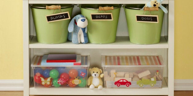 Interior of a child's play room showcasing a white shelf photographed on a wood laminate floor against a yellow painted wall with two hanging blank canvas that have paths for dropping in other art work. White shelf suggests storage ideas, it has three chalk labeled buckets that can store various kid items and two plastic containers with lids one has blocks and the other plastic balls.
