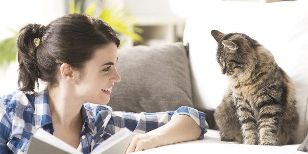 Smiling woman playing with her cat and holding a book in the living room.