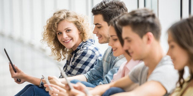 College friends sitting on the floor and watching pics on their gadgets at break. Side view.